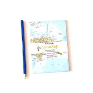 Könnte beinhalten: Ein Notizbuch mit einer Karte der Dominikanischen Republik, mit dem Text "Notizbuch" und "DomRep". Ein blauer Bleistift liegt neben dem Notizbuch. Das Notizbuch hat ein weißes Band mit Text und einem Palmenlogo.