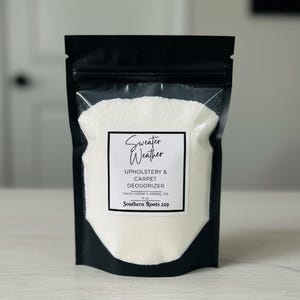 May include: A black pouch with a clear window containing white powder. A white label reads "Sweater Weather Upholstery & Carpet Deodorizer" with the text "Southern Roots 229" below. The product is hand crafted in Albany, GA.