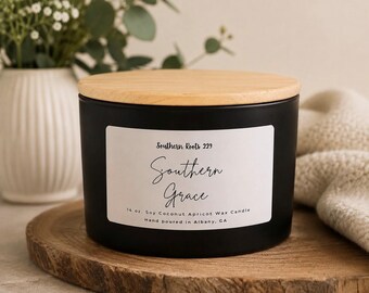May include: A matte black candle jar with a light wood lid. The white label features the text Southern Roots 229, Southern Grace, and 14 oz. Soy Coconut Apricot Wax Candle, Hand poured in Albany, GA. The container has a cylindrical shape.