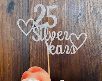 May include: A white cake with gold leaf accents, topped with two white meringue cookies, a peach, and a silver glitter cake topper that reads "25 Silver Years" with two hearts.