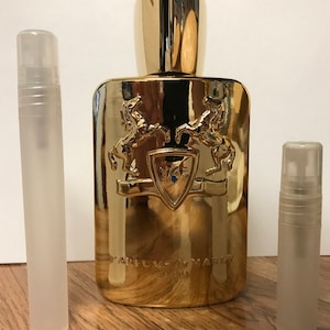 May include: A gold bottle of perfume with a gold cap and a gold label that says "Parfums de Marly" and "1743". The bottle is surrounded by two clear glass perfume sample vials.