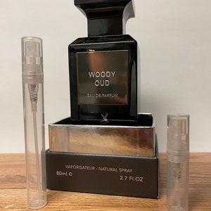 May include: A black bottle of Woody Oud Eau de Parfum with a silver label. The bottle is sitting on a brown wooden surface with two clear glass sample bottles.