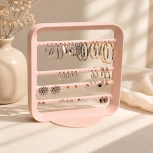 May include: A light pink, square-shaped jewelry organizer stand with four horizontal bars featuring evenly spaced holes for hanging earrings. The stand is constructed with a sturdy base and holds various silver and gold-toned hoop and stud earrings.