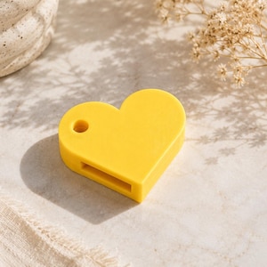 May include: A bright yellow heart-shaped object with a smooth, matte finish. The item features a small circular hole near the top left lobe and a rectangular slot along the bottom edge, providing a functional design for attachment or cord threading.