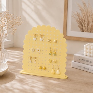 Puede incluir: Un expositor de joyas de color amarillo pálido, con forma de arco, bordes festoneados y una rejilla perforada. El soporte sostiene varios pares de pendientes, incluyendo formas de corazón, flor, lazo y lágrima, sobre una base rectangular.