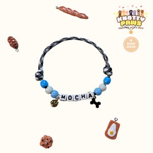 May include: A handmade pet collar with the name "MOCHA" spelled out in white letter beads. The collar features blue and white beads, a black and white rope design, and charms including a bone and a paw print. The image also includes food-themed charms.