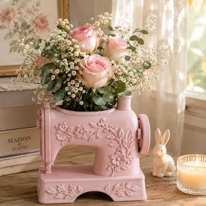 Puede incluir: Un jarrón decorativo de color rosa pastel con forma de máquina de coser vintage, que presenta detalles florales en relieve en el lateral y la base. El jarrón está lleno de un ramo de rosas de color rosa claro, paniculata blanca y hojas verdes.