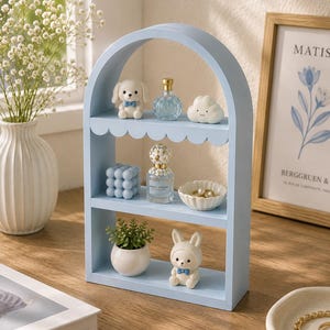 May include: A light blue arched wooden shelf with three tiers. The top shelf holds a white dog figurine, a small blue glass perfume bottle, and a white cloud figurine. The middle shelf features a blue bubble candle, a perfume bottle, and a small white bowl with rings. The bottom shelf contains a small potted green plant and a white rabbit figurine.