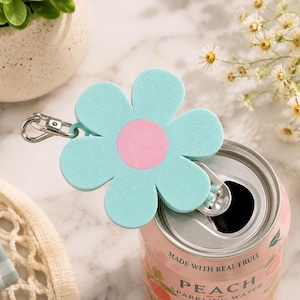 May include: A light blue flower-shaped soda can opener with a pink center and a silver metal ring attachment. The opener is positioned on top of a pink aluminum can labeled Peach Sparkling Water, which features text reading Made with real fruit.