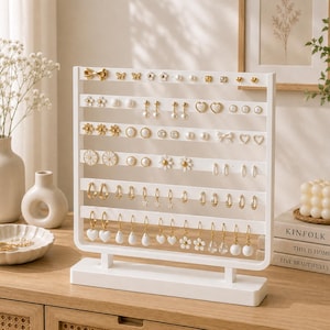 Puede incluir: Un expositor de pendientes rectangular de color blanco con varias barras horizontales que tienen orificios perforados. El soporte contiene una variedad de pendientes dorados y blancos, incluyendo tachuelas, aros y colgantes con formas de flores, corazones y lazos.