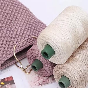 May include: Close-up of three spools of thread in cream, mauve, and tan colors. The spools have green cores and are positioned near a mauve crocheted bag. The thread appears to be a smooth, shiny material, suitable for various crafting projects.