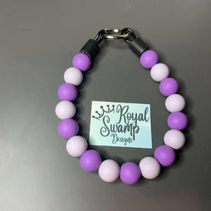 May include: A necklace made of alternating purple and lilac beads, with a black clasp and connectors. The necklace is displayed with a card that reads "Royal Swamp Designs".