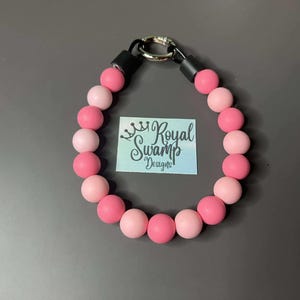 May include: A necklace made of pink and pale pink beads, with a black clasp and black detailing. The necklace has a tag in the centre with the text "Royal Swamp Designs".