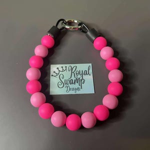 May include: A beaded dog collar featuring alternating pink and hot pink beads. The collar has a black clasp and a silver metal ring. A small tag with the text "Royal Swamp Designs" is visible.