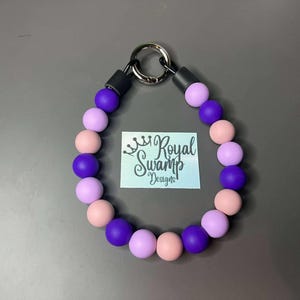 May include: A beaded collar with a silver ring and black details. The collar has round beads in shades of purple and pink. A small tag with the text "Royal Swamp Designs" is in the centre.