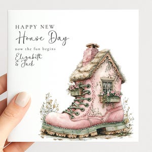 May include: A square greeting card featuring a pink boot-shaped house with a thatched roof, chimney, and window boxes filled with flowers. The text reads: HAPPY NEW House Day, now the fun begins, Elizabeth & Jack. The design includes green laces.
