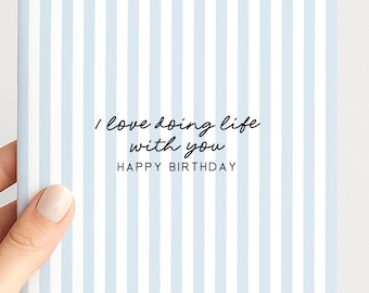 May include: A square greeting card featuring a pattern of vertical light blue and white stripes. The text "I love doing life with you" is printed in a black script font, followed by the words "HAPPY BIRTHDAY" in a simple black sans-serif font.