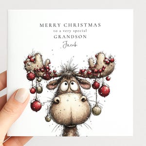 May include: A square white greeting card featuring a cartoon moose with wide eyes and large antlers decorated with hanging red and gold Christmas ornaments. The text reads MERRY CHRISTMAS TO A VERY SPECIAL GRANDSON Jacob in black font.