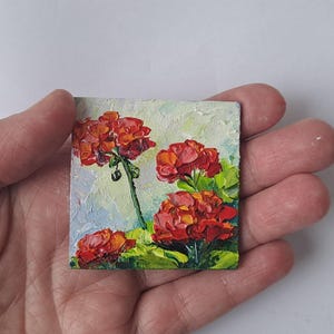 May include: A small square oil painting of vibrant red geranium flowers with green leaves and stems. The artwork has a textured, impressionistic style, set against a light blue and white background. The painting is held in a hand.