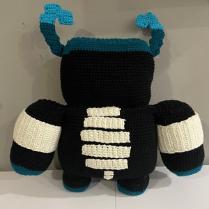 May include: Black crocheted plush toy with white crocheted details resembling a skeleton. The toy has teal blue crocheted arms and legs.