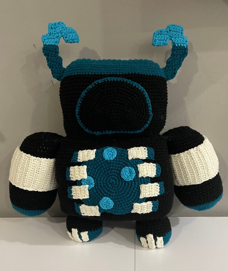 May include: A crocheted robot toy with black, white, and teal blue colors. The robot has two arms, two legs, and a round head with a teal blue face.
