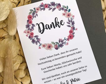 May include: A white card with the word "Danke" in black script, surrounded by a floral wreath of pink and purple flowers. The card includes additional text in German and is placed on a woven background.