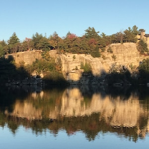 May include: A scenic view of a lake reflecting the rocky cliffs and trees on the opposite shore. The water is calm and still, creating a mirror-like reflection of the surrounding landscape.