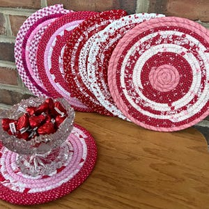 May include: A set of six red and white fabric coasters with a floral pattern. The coasters are arranged in a circle on a wooden table. A glass bowl filled with red candy hearts sits on a larger red and white fabric coaster.