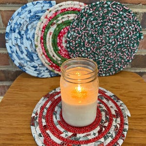 May include: Four round, crocheted, fabric trivets in blue, red, green, and white patterns. A clear glass jar with a white candle inside sits on the red and white trivet.