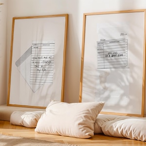 May include: Two framed art prints with wooden frames. One print features the text "you met me at a very strange time in my life." The other print has the text "get lost in a good plot." Beige pillows and a wooden vase are in the foreground.