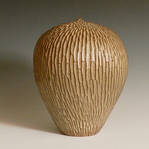 Könnte beinhalten: Eine beige Keramikvase mit einem strukturierten, vertikal gerillten Muster. Die Vase hat einen abgerundeten Körper, der sich an der Basis verjüngt, und eine kleine, strukturierte Oberseite. Die Vase ist etwa 30 cm hoch.