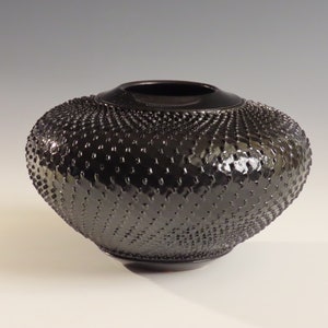 Puede incluir: Un jarrón de cerámica negro con una superficie texturizada y con hoyuelos. El jarrón tiene una forma redondeada, ligeramente ovalada.