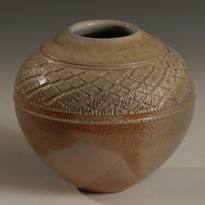 Könnte beinhalten: Eine runde, beige Keramikvase mit einem strukturierten, gewebten Design um die Mitte. Die Vase hat eine leicht cremefarbene Glasur mit einem dezenten Braunton.