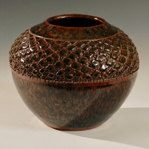 Könnte beinhalten: Eine handgefertigte Keramikvase mit einem strukturierten, rautenförmigen oberen Bereich und einem glatten, abgerundeten Boden. Die Vase hat eine satte, erdige Farbpalette aus Braun, Grün und Rotbraun, die eine rustikale Ästhetik erzeugt.