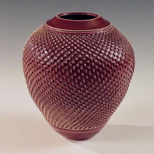 Op de afbeelding: Een diep rode keramische vaas met een getextureerd, ruitvormig patroon. De vaas heeft een rond lichaam en een licht uitlopende opening.
