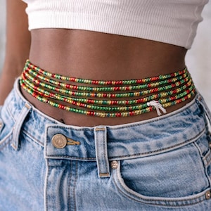 Könnte beinhalten: Taillenperlen in Rot, Grün und Gold sind um die Taille gewickelt. Die Perlen werden über einer blauen Jeans und einem weißen Crop-Top getragen. Die Perlen sind mit einer weißen Schnur zusammengebunden.