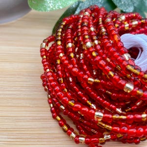 Op de afbeelding: Een close-up van een rode en gouden kralen armband. De kralen zijn klein en rond en zijn gerangschikt in een spiraalvorm. De armband is gemaakt van veel strengen kralen en is erg lang.