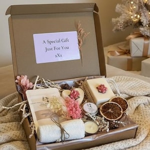 Può includere: Una confezione regalo aperta con saponi, candele e fette d'arancia essiccate. La scatola è appoggiata su una coperta color crema. Un albero di Natale con decorazioni dorate e regali confezionati sullo sfondo.