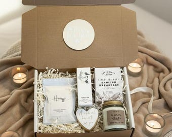 Puede incluir: Una caja de regalo abierta con una vela, té y una galleta en forma de corazón. La caja está rodeada de velas encendidas y una manta suave. La caja tiene un letrero redondo de madera que dice "Lo mejor está por venir."