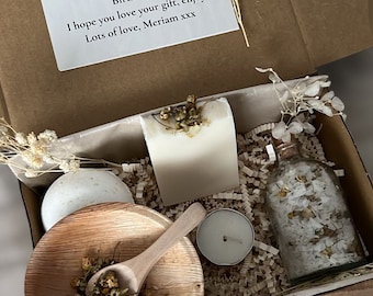 Könnte beinhalten: Eine Geschenkbox gefüllt mit Selbstpflegeartikeln, darunter eine weiße Seife, eine kleine Glasflasche gefüllt mit weißen Badesalzen, eine Holzschale mit getrockneten Blumen, eine kleine weiße Kerze und ein weißes Handtuch, das mit Schnur gebunden ist. Die Box ist mit braunen Papierschnitzeln ausgekleidet.
