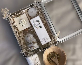 Könnte beinhalten: Eine silberne Metallbox gefüllt mit Bad- und Körperpflegeprodukten. Die Box enthält eine weiße Seife mit einer Blume darauf, eine klare Glasflasche mit weißen Badesalzen, eine weiße Flasche After-Oil, eine kleine weiße Kerze und eine kleine Schale mit getrockneten Kamillenblüten. Die Box steht auf einer hellbraunen Oberfläche.