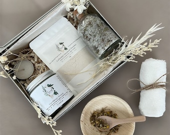 Könnte beinhalten: Eine silberne Metall-Geschenkbox gefüllt mit Bad- und Körperpflegeprodukten. Die Box enthält ein Glas Caribbean Dream Body Scrub, einen Beutel mit kolloidalem Haferflockenpulver, eine Glasflasche mit Badesalz, eine kleine weiße Kerze und ein weißes Waschlappen. Die Box ist von getrockneten Blumen umgeben.