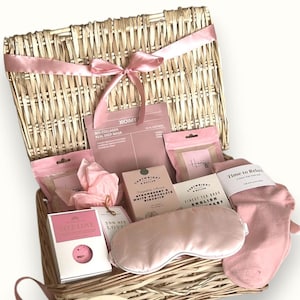 Pode incluir: Uma cesta de vime cheia de itens de autocuidado rosa e branco, incluindo uma máscara para dormir, bombas de banho, saquinhos de chá e uma máscara facial. A cesta é amarrada com uma fita rosa e tem uma etiqueta de "Feliz Aniversário".