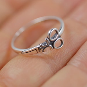 May include: A silver ring with a scissors design. The ring features a detailed scissor blade and handle, with the scissor blades forming two circles. The ring is shown on a person's hand.
