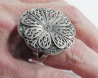 May include: A large, silver-toned ring with an intricate floral design. The ring features a detailed, openwork pattern with a central flower-like element and a beaded border. The ring is worn on a finger, showcasing its size and ornate style.