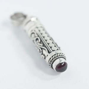 May include: A silver pendant with detailed scrollwork and a cylindrical shape. It has a small, round, dark red gemstone at the base. The top has a loop for a chain. The pendant is set against a white background.