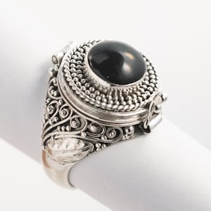 May include: A silver ring with an ornate design and a large, round, black stone. The ring features detailed filigree and beaded accents around the stone. The ring is displayed on a white surface.