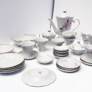 May include: A white porcelain tea set with a floral design. The set includes a teapot, cups, saucers, sugar bowl, creamer, and serving dishes. Each piece is decorated with pink flowers and gold trim. The set is arranged on a white surface.