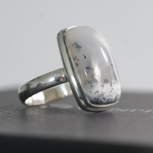 May include: A silver ring featuring a large, rectangular dendritic agate stone. The stone is primarily white with delicate black and gray patterns. The ring has a polished silver band and bezel setting, showcasing the natural beauty of the gemstone.