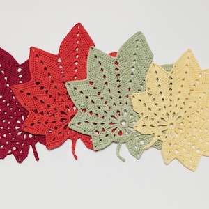 May include: A collection of five crocheted leaf-shaped placemats in shades of red, green, and yellow. The leaves have intricate openwork patterns and are arranged in a row, showcasing their autumnal colors and textures.